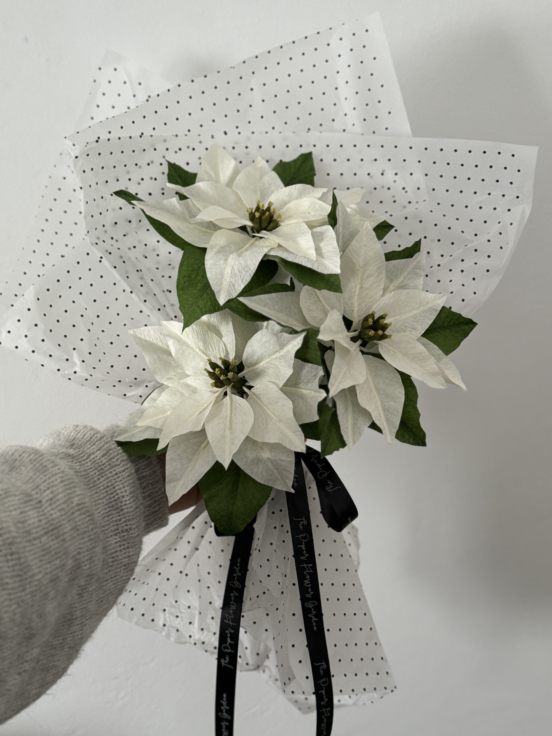 Ramo de tres Flores de Pascua blancas hechas a mano con papel crepé y hojas verdes, envuelto en papel de lunares.